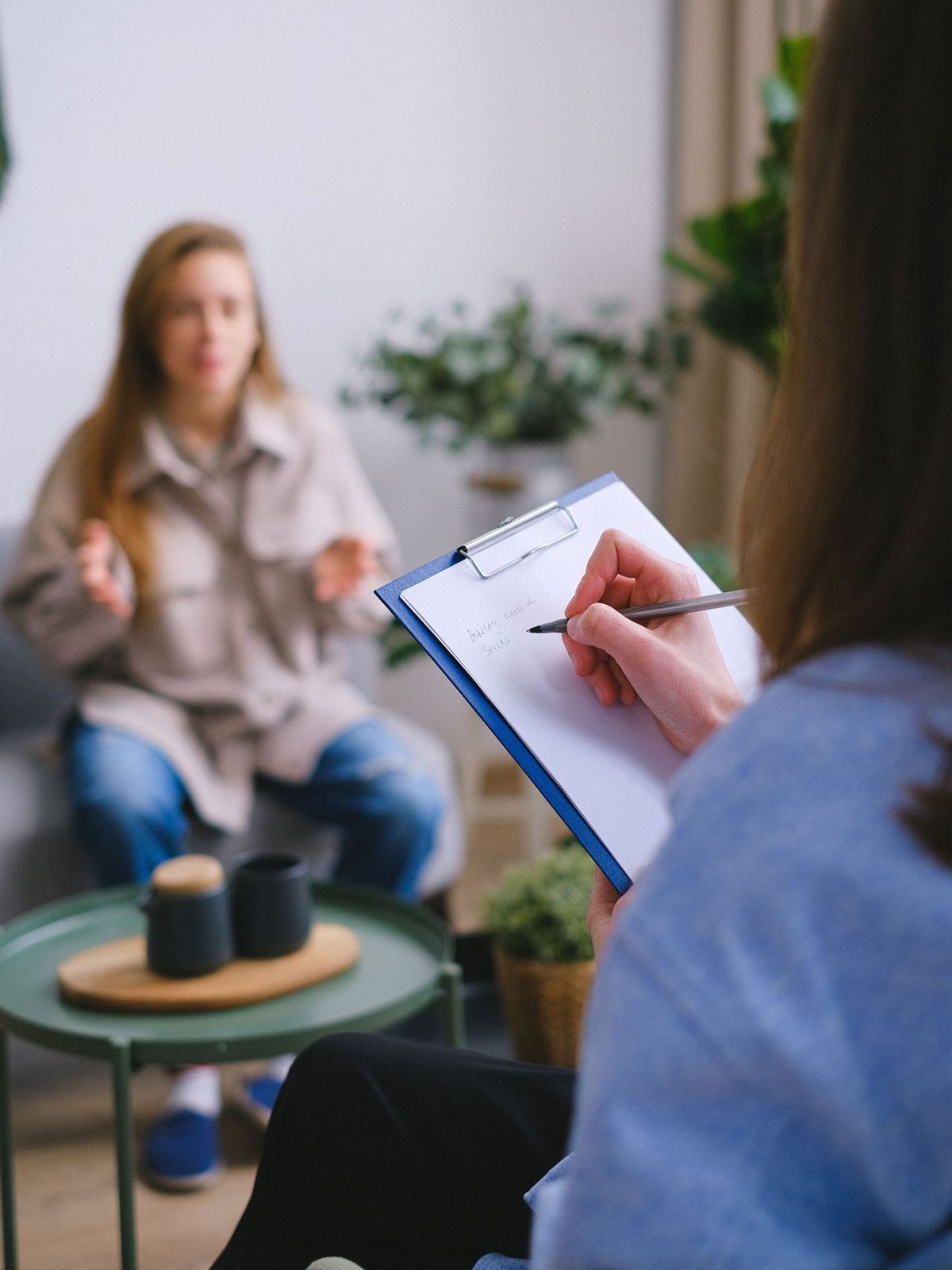 A clinician making notes on a clipboard while a patient talks, in a room with plants and soft daylight.