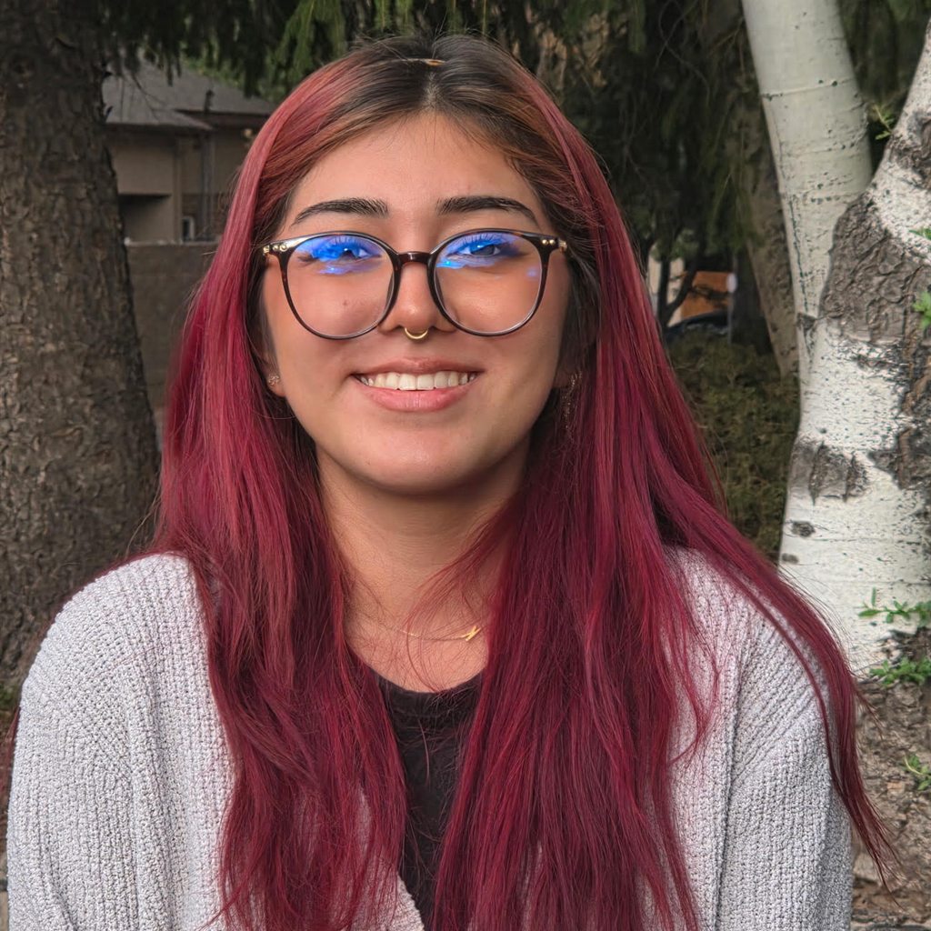 Kaileena Riley, smiling, wearing glasses and a grey cardigan, photographed outdoors in front of an aspen tree.
