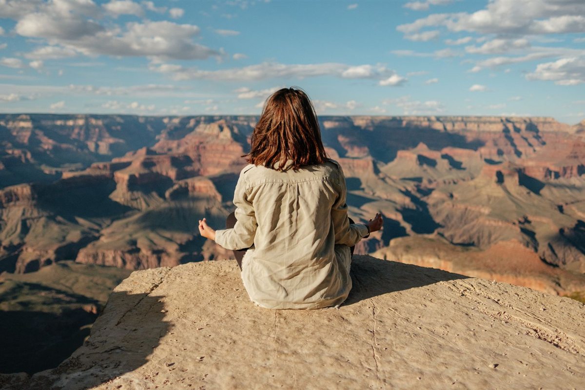A woman sitting cross-legged on a rock ledge above the Grand Canyon, seen from behind, looking out over the canyon in late afternoon light.