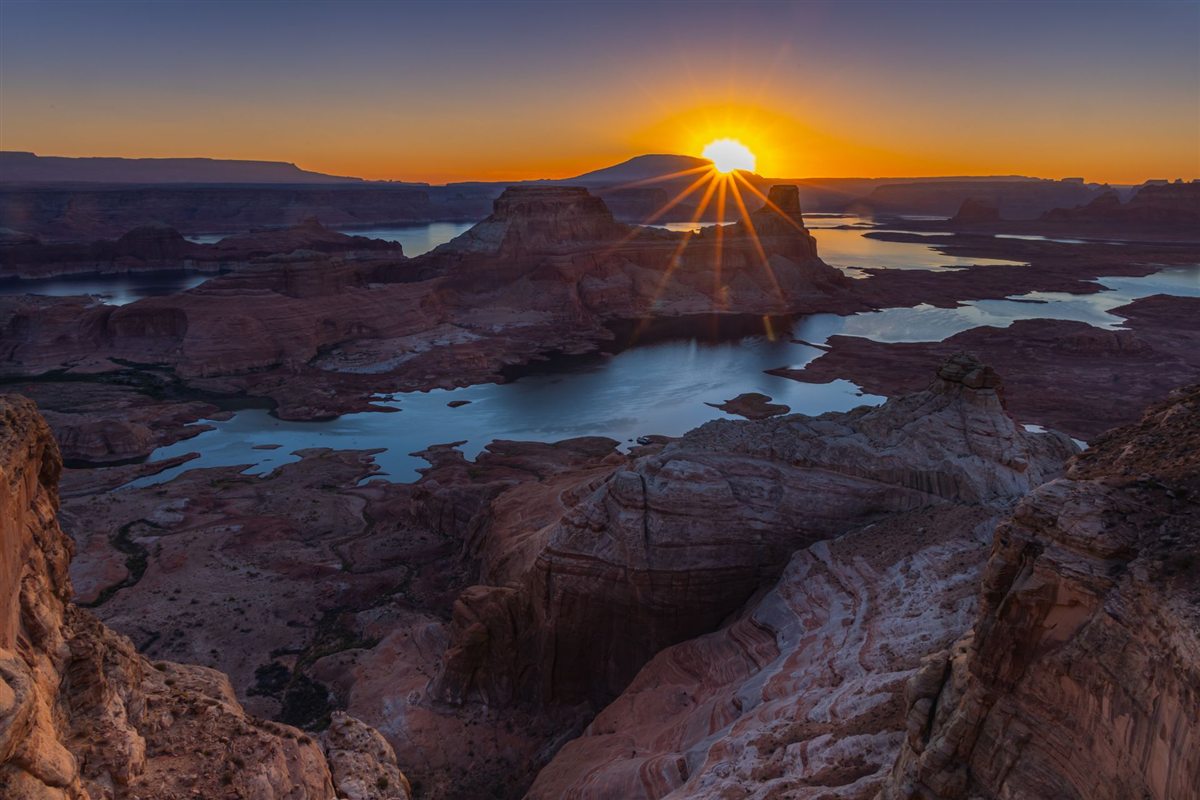 Sunrise over Lake Powell, the sun breaking in a starburst above a butte with the lake and canyon country spread out below.
