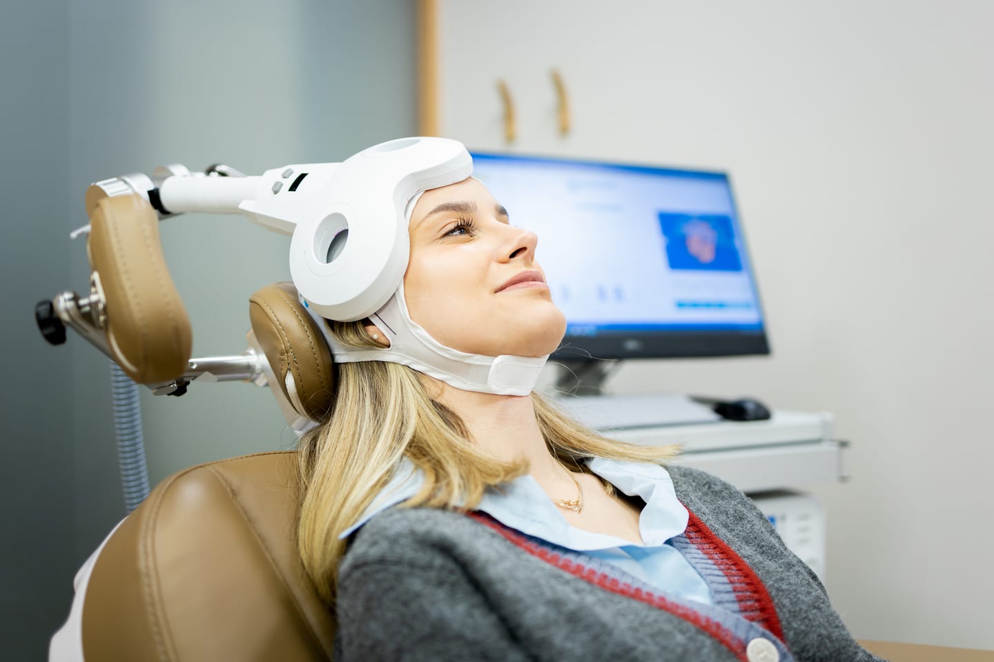 A smiling woman reclines in a treatment chair, a white TMS coil on an adjustable arm strapped against the front of her head.